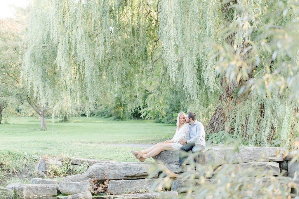 couple sitting on the rocks by the water in front of the willow tree at dominion arboretum