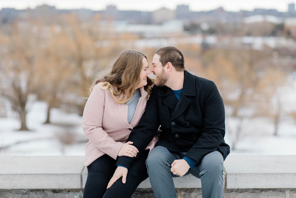 couple sitting beside each other nose to nose sitting on a rock ledge at winter engagement session