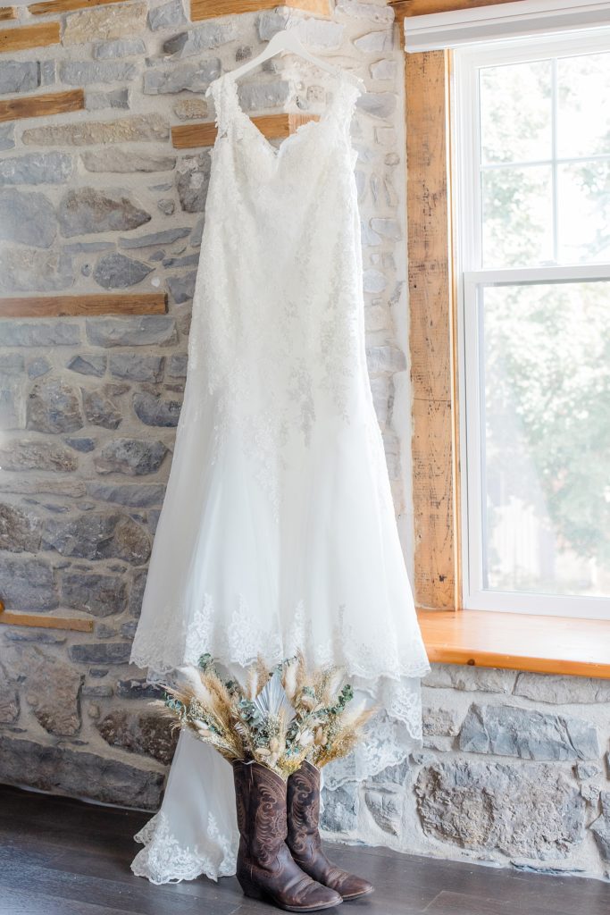 renewed with love wedding dress hanging up on stone wall with cowboy boots below it and dried florals sticking out of cowboy boots for country themed wedding in ottawa