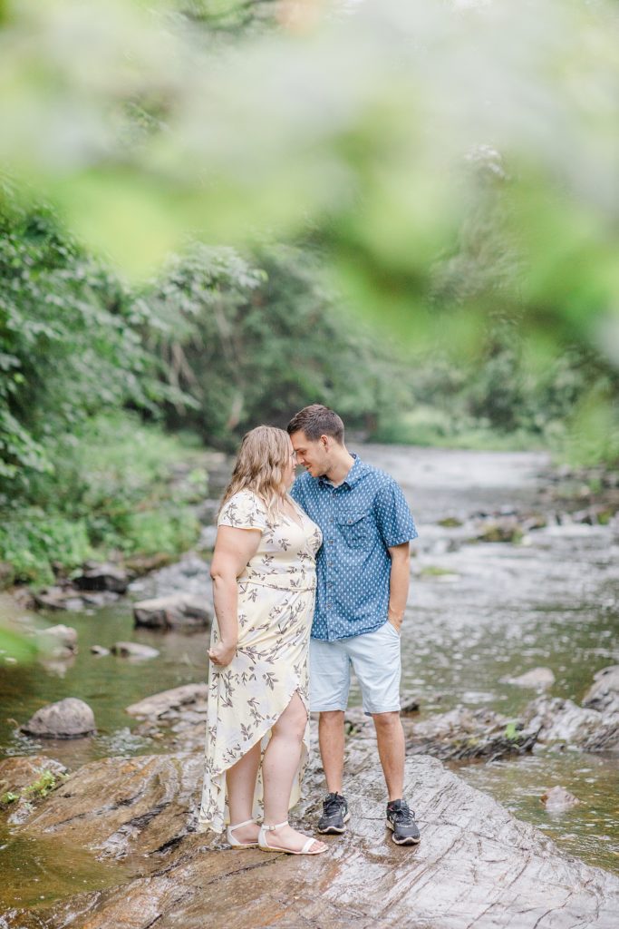 couple standing on rock together in the middle of the water at rainy summer Mill of Kintail Engagement session with Brittany Navin Photography in Almonte, ON
