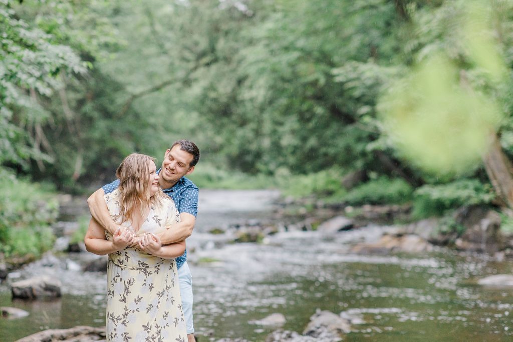 he has his arms wrapped around her as they stand on a rock in the middle of the water during rainy summer engagement session in Almonte, ON, with Brittany Navin Photography