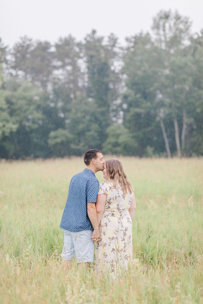 He is leaning over to giver her a foehead kiss in the rain in the field during Almonte engagement session with Brittany Navin Photography
