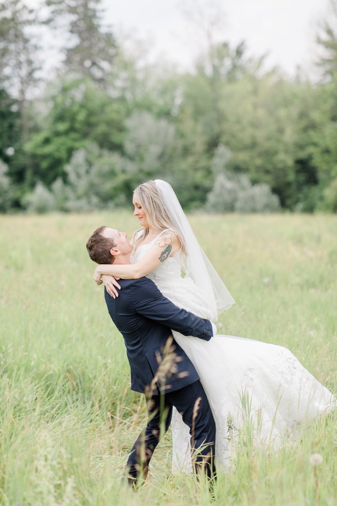 groom picking up bride in the middle of long grassy field at Mill of Kintail Wedding with Brittany Navin Photography in Almonte, Ontario