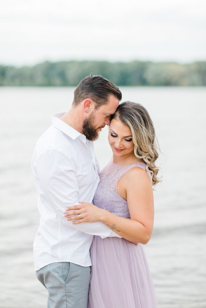 she is looking down over her shoulder as her fiance rests his head gently on hers during Petrie Island Beach Engagement session