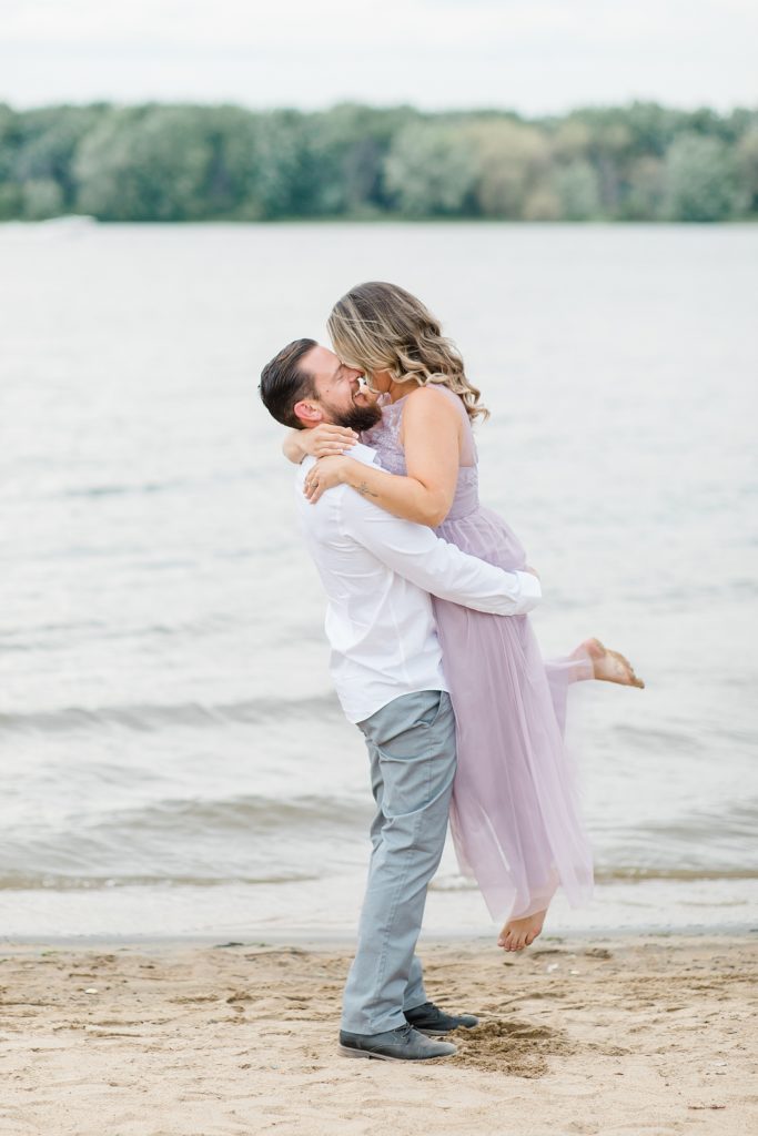 he is lifting his fiance up during Petrie Island Beach Engagement session