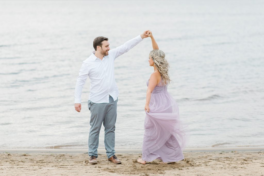 fiances dancing on the beach in ottawa during their engagement session wearing a lavender maxi dress and white button down with grey dress pants