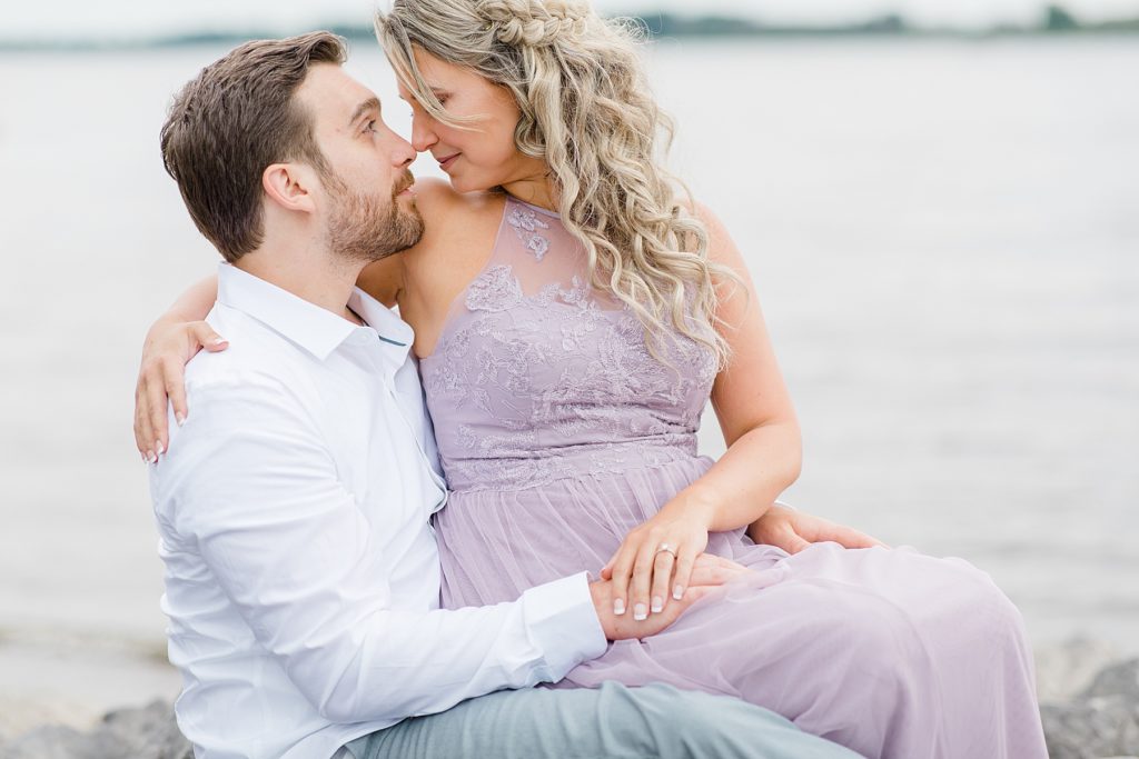 bride to be sitting on fiance's lap on the rocks along the beach during ottawa engagement
