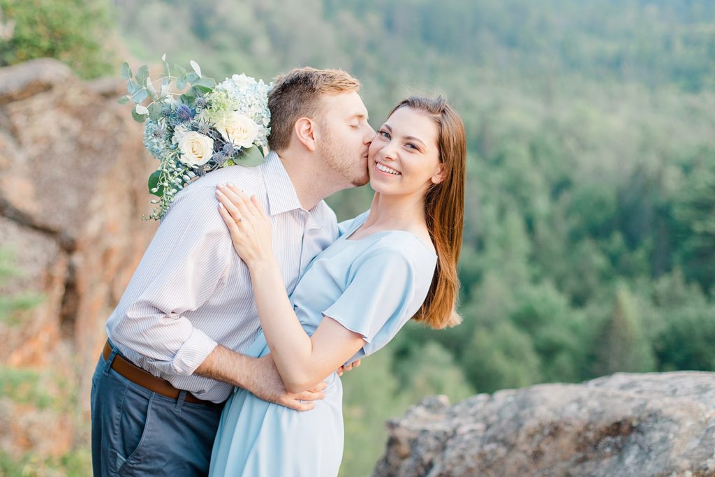he is giving her a kiss on the cheek while she smiles at the camera at Eagle's Nest Engagement in Calabogie, Ontario with Brittany Navin Photography