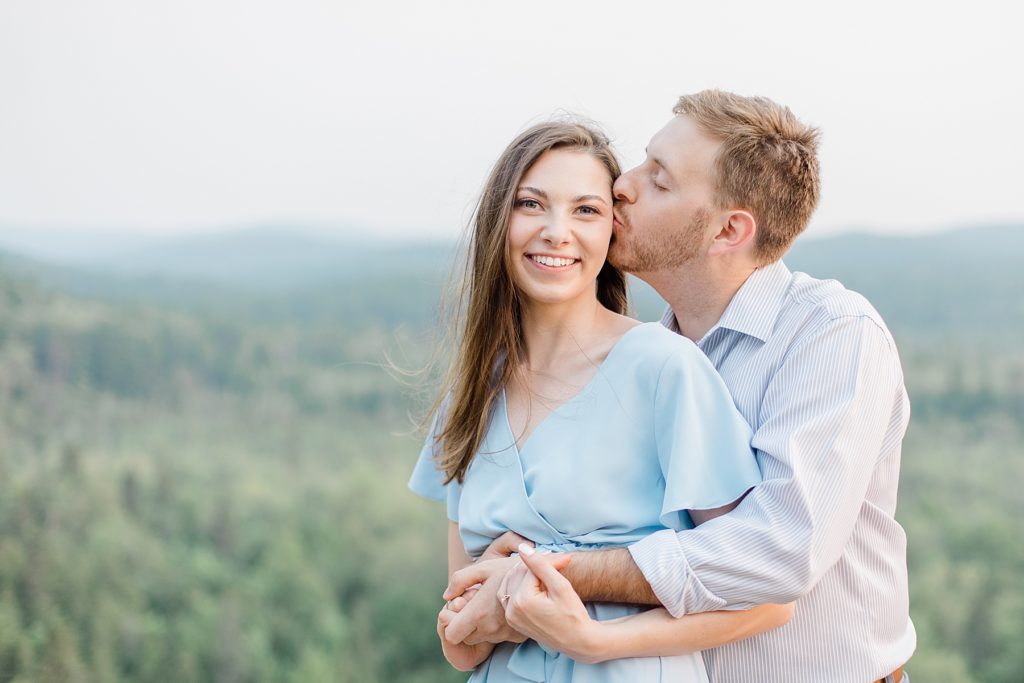 he is giving her a kiss on the cheek as she smiles at the camera at Eagle's Nest Engagement in Calabogie, Ontario with Brittany Navin Photography