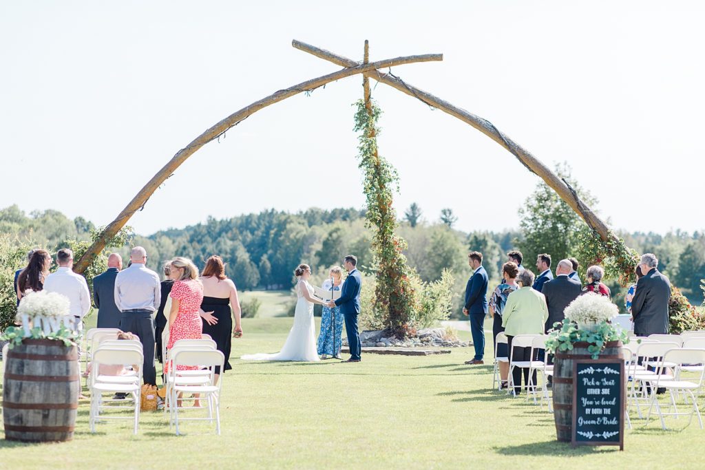 wedding ceremony under giant wooden arch at timber run golf course wedding in Lanark, Ontario photographed by Brittany Navin. Altar is in ideal lighting for ceremony.