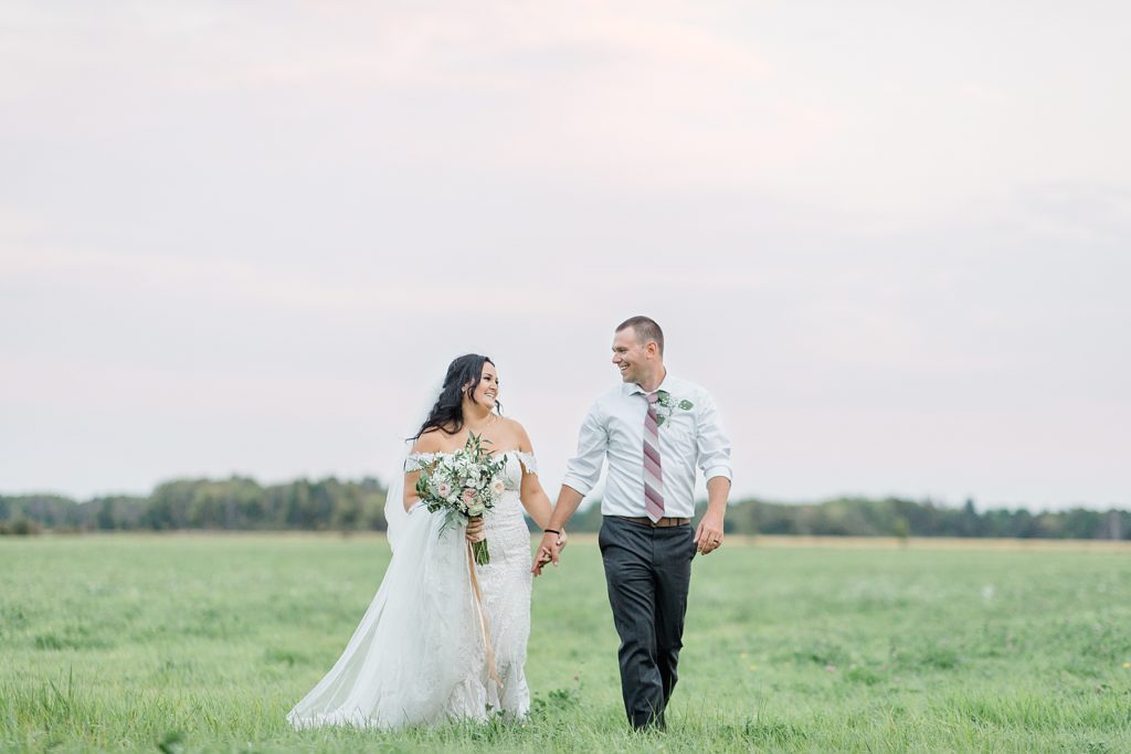 bride and groom walking through field with a faint pink sky at Adelina Barn Wedding Photographed By Brittany Navin Photography