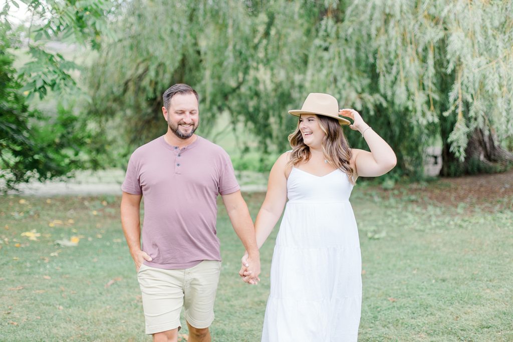 couple walking together as she holds her hat in the wind at dominion arboretum engagement session phtoographed by Brittany Navin Photography