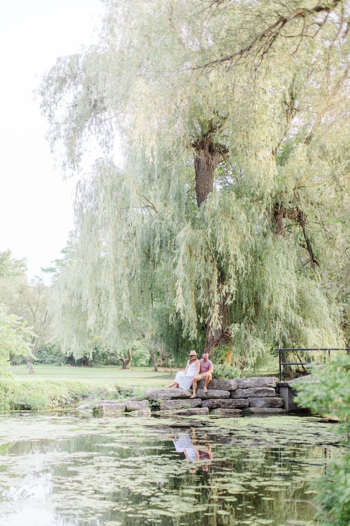 Couple sitting together on rocks by little creek with willow tree behind them at dominion arboretum engagement session phtoographed by Brittany Navin Photography