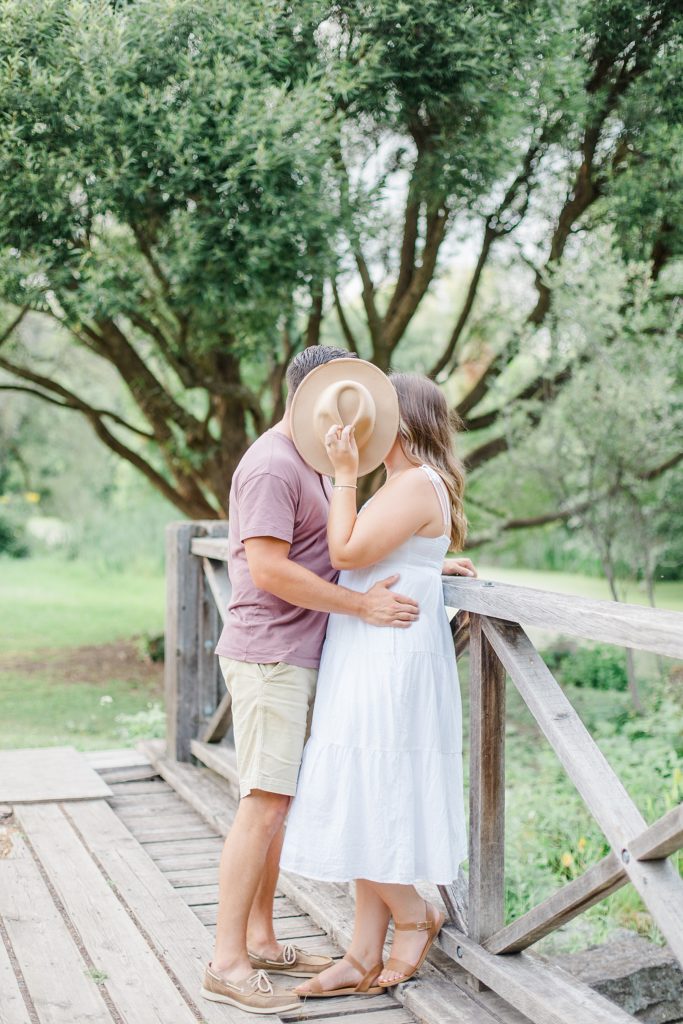 couple hanging out on a bridge with her hat covering their faces as they go in for a kiss at dominion arboretum engagement session phtoographed by Brittany Navin Photography