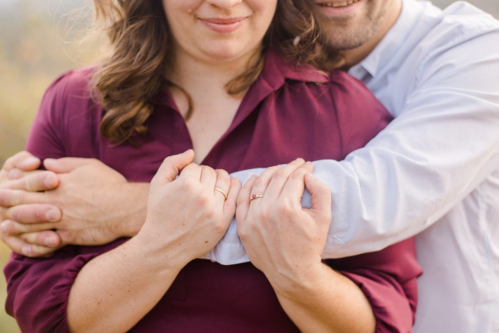 close up of her engagement ring on her hands holding his arm thats wrapped around her photographed by Brittany Navin Photography