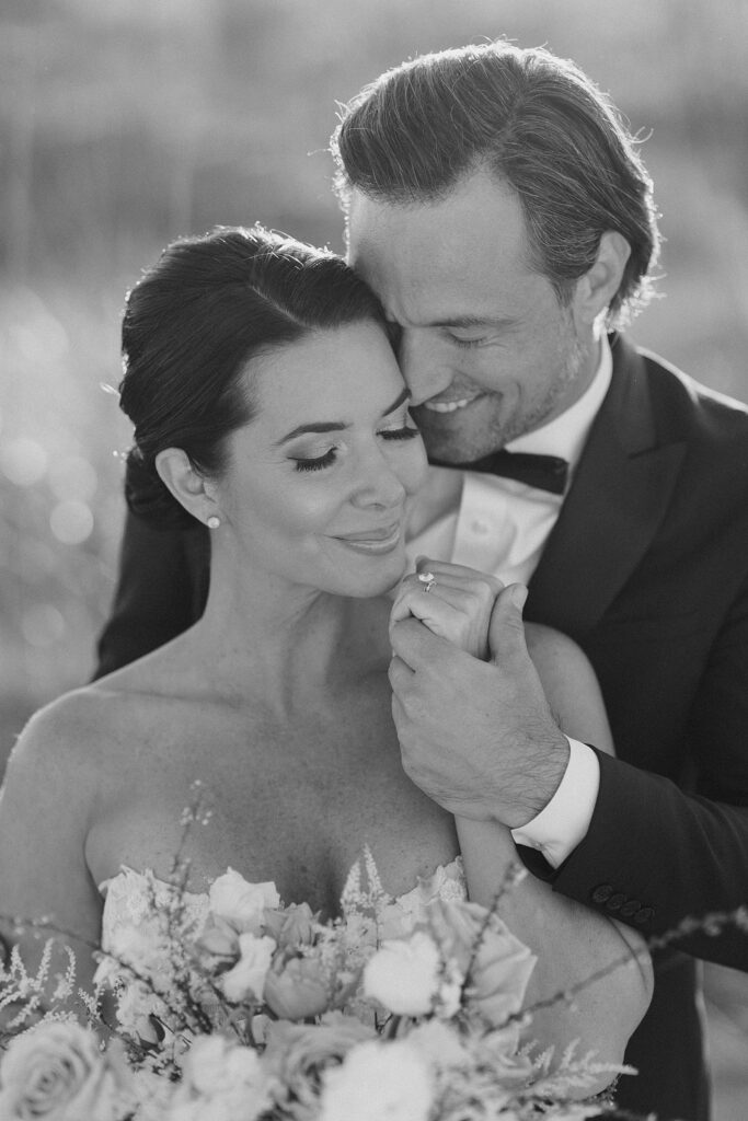 black and white image of groom smiling and nuzzling into his bride as he pulls up her hand with her ring for a kiss at destination wedding photographed by Brittany Navin Photography
