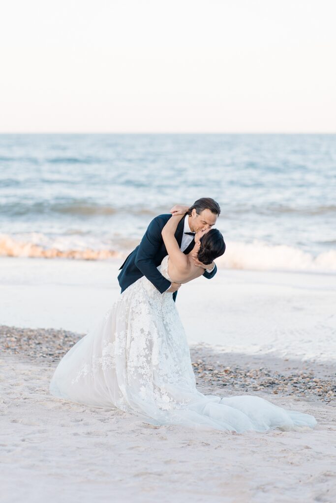 husband dips his wife for a kiss during wedding portraits on the beach at the coastal opulence beach wedding editorial photographed by Brittany Navin Photography