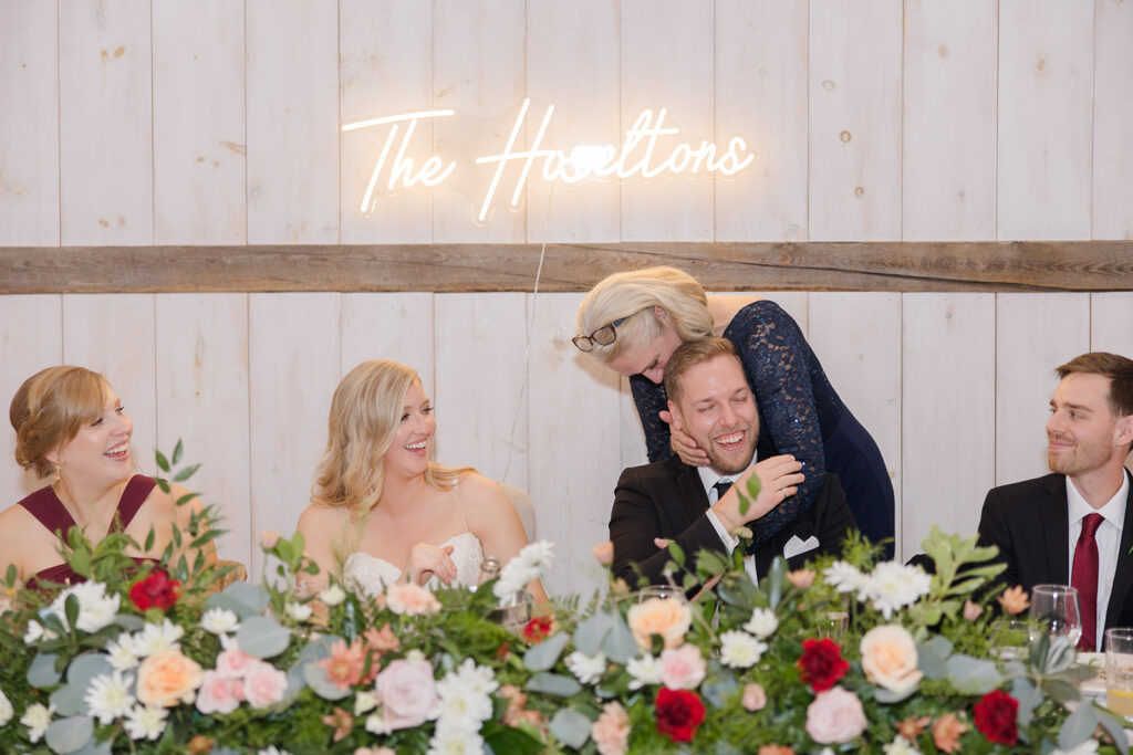 grooms mom attacks son with hugs while hes at his seat during reception at Stonefields Estate wedding photographed by Ottawa Wedding Photographer Brittany Navin Photography