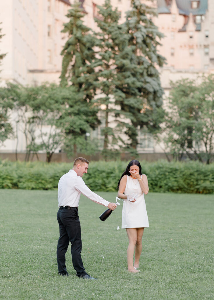 Couple popping a champagne bottle on the lawn near the Château Laurier during their playful summer engagement session.