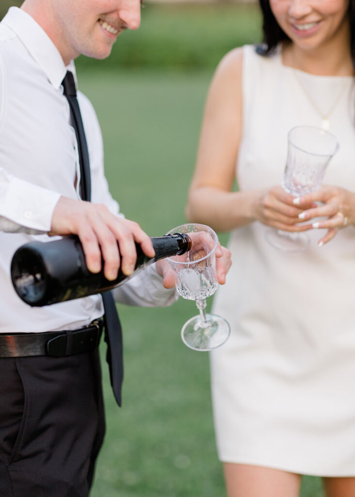 Couple popping a champagne bottle on the lawn near the Château Laurier during their playful summer engagement session.