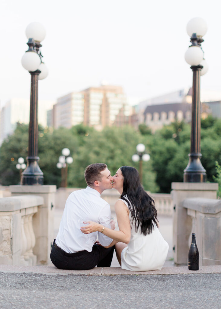 Couple popping a champagne bottle on the lawn near the Château Laurier during their playful summer engagement session.